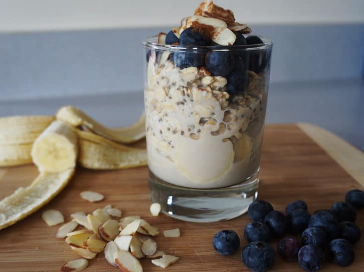 Wooden board topped with banana, blueberries, sliced almonds and cup. Cup contains quick oats topped with fruit and nuts.