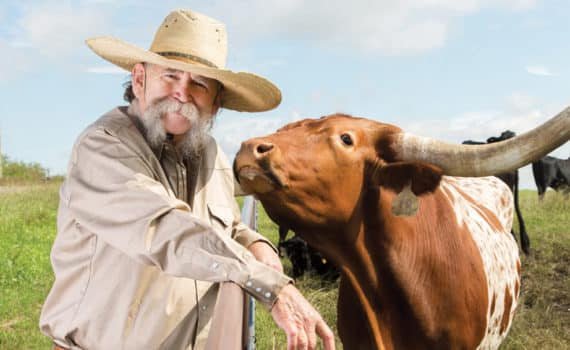 Man in cowboy hat and large mustache standing next to fence and a bull on the other, whose face is close to the man's.