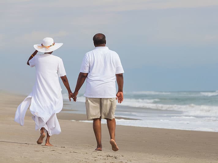 Man and woman holding hands on a beach wearing white and walking away. Woman is wearing a white sun hat.