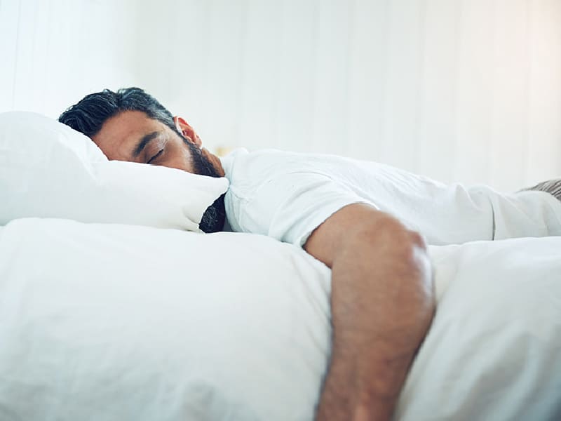 Man with dark hair and beard asleep on a bed with white sheets and pillow cases. His arm is dangling off the side of the bed.