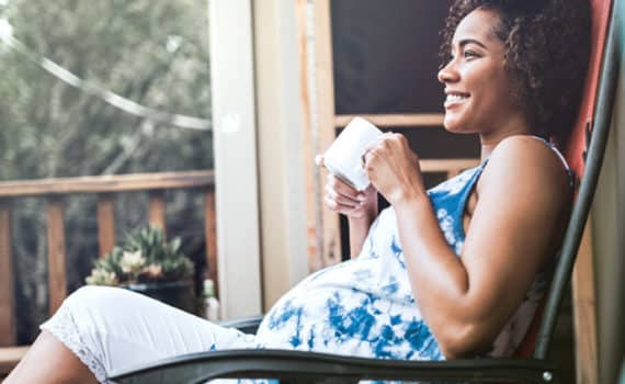 Pregnant woman sitting in rocking chair on a porch with a mug in her hand.