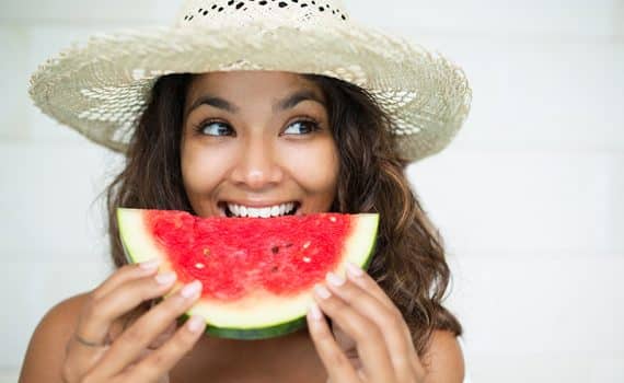 Woman with brown hair wearing straw hat eating a half-moon slice of watermelon.