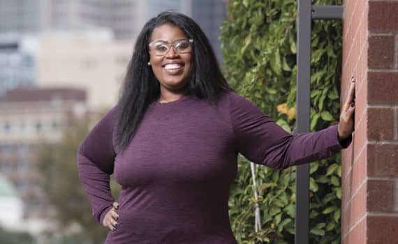 LaRonda Darby after bariatric surgery, smiling with hand on hip leaning on a wall with ivy with city background.