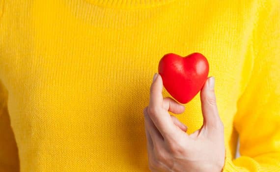 Woman holding a red heart next to a yellow sweater; women and heart attack symptoms.