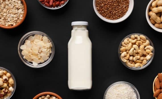 bottle of plant-based milk surrounded by bowls of nuts, rice, and oats