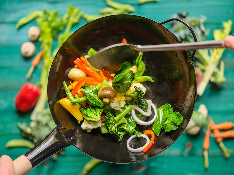 A variety of fresh market vegetables including broccoli, carrot, capsicum, snow peas, red onion, green beans, celery and mushrooms being stir-fried and cooked in a hot wok, with an out of focus turquoise-colored wooden, vegetable covered table in the background.