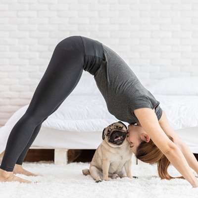 Woman in black workout attire doing the downward dog, a yoga position, while her pug sits under her, promoting self-care during the pandemic
