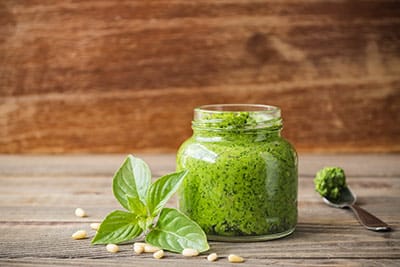 Glass jar with pesto inside, sitting on a wooden surface with basil leaves and pine nuts, two other Italian foods