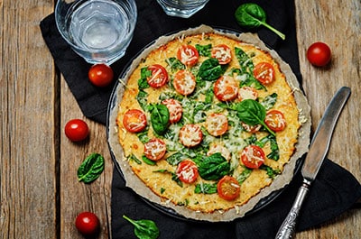 Cauliflower pizza with tomatoes and spinach, sitting on wooden tabletop with knife and glass table setting