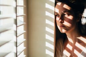 Woman looking outside, with the blinds casting shadows on her face and body