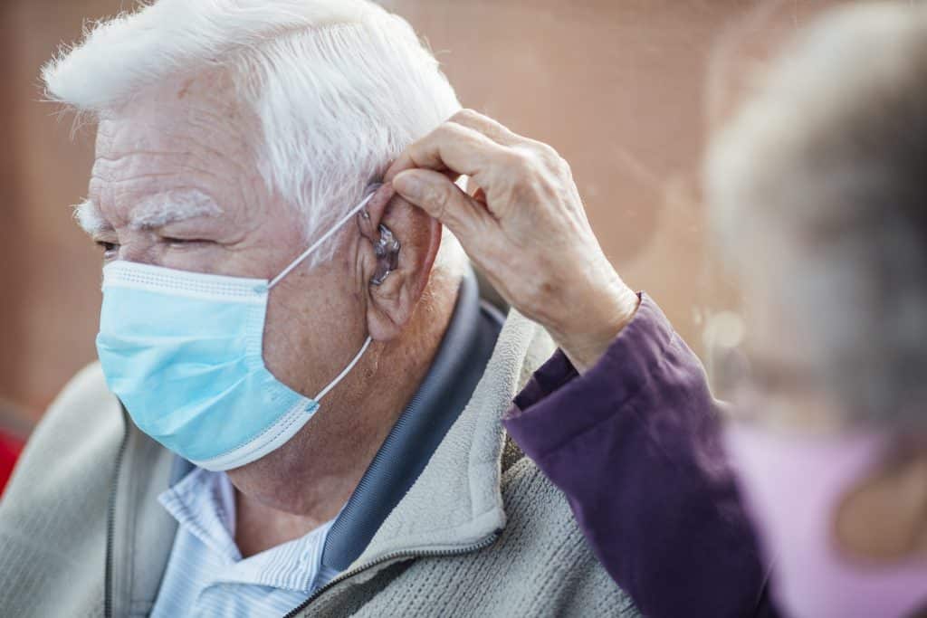 Senior helping other senior attach his mask strap around his ear with a hearing aid