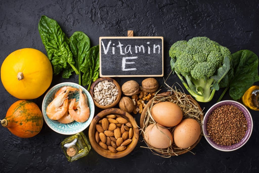 Chalkboard with "vitamin E" written on it, surrounded by foods such as squash, shrimp, nuts, eggs, seeds, oil, broccoli, and more.