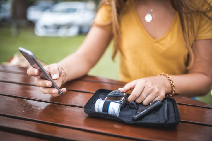 Woman in yellow shirt reading from a phone and holding a bag with a blood sugar monitor and other related items