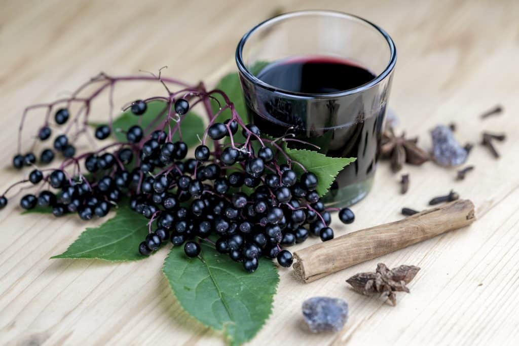 Raw elderberries sitting on a wood table next to a glass of elderberry juice