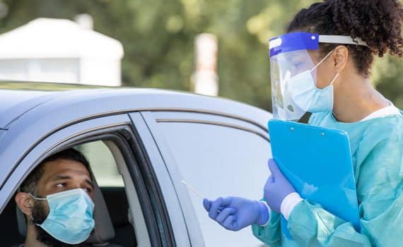 Man with medical face mask sitting in a car while getting ready to receive a COVID-19 test from a medical professional in blue scrubs and a face shield