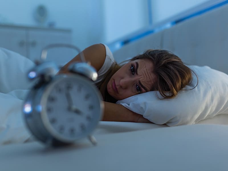 Woman in bed looking at alarm clock, used to explain sleep aids pros and cons
