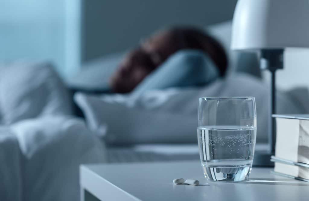 A glass of water and two pills sit on a nightstand next to a person sleeping in bed