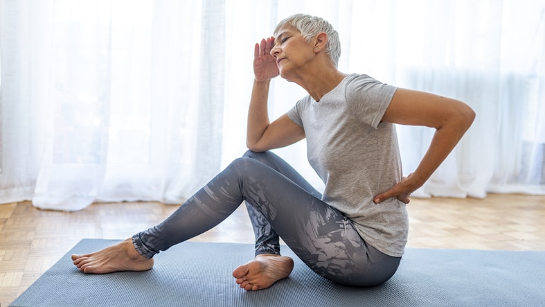 An older woman sitting on a yoga mat, holding her head and lower back 