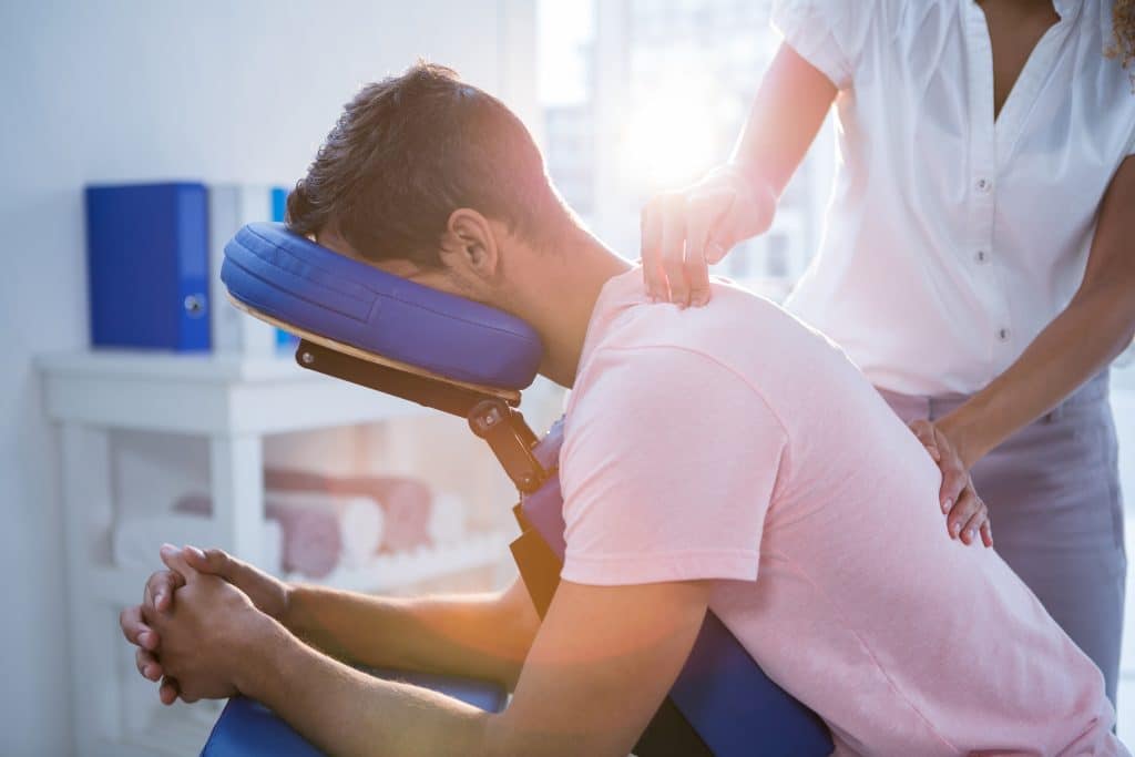 A person with their face in a frontward-facing orthopedic chair while a specialist puts pressure on the upper back