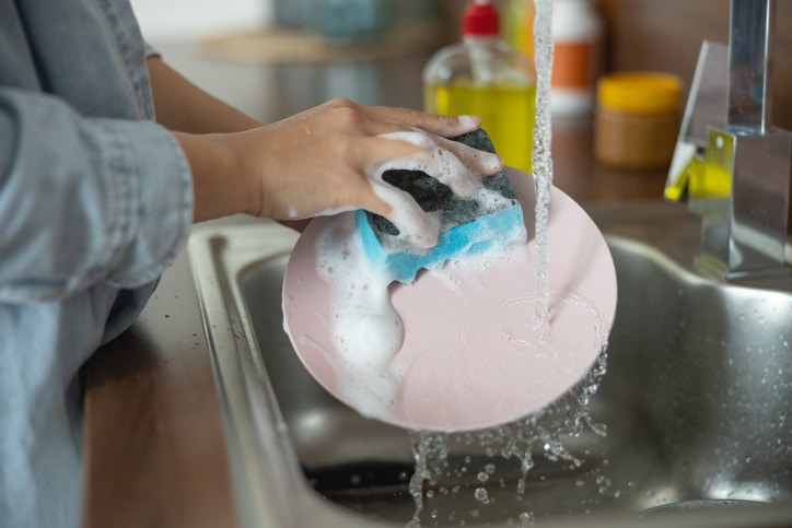 A person cleaning dishes with a blue soapy sponge under running water