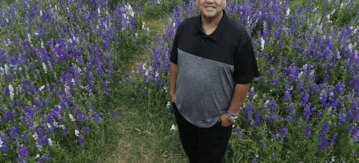 Daniel Lauriano smiling at the camera and standing in a field of purple flowers,photographed after his widow maker heart attack