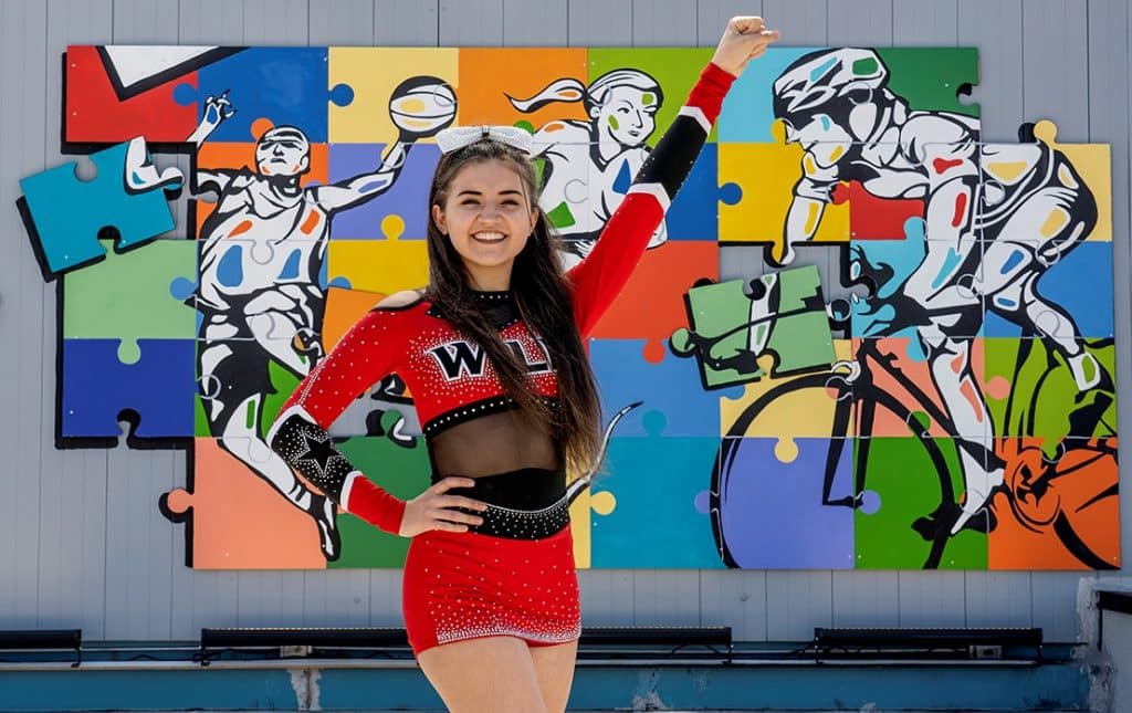 Chloe Galvez dressed in her cheerleading uniform in front of a mural with puzzle pieces and bright colors