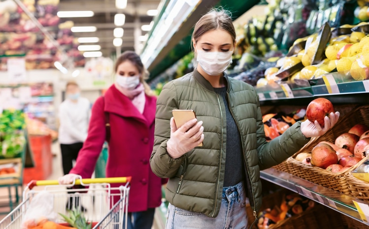 Two women wearing masks while at the grocery store and holding items in the produce section