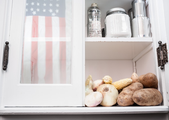 A cabinet other with potatoes, corn, and onions on one shelf and jars on the top shelf