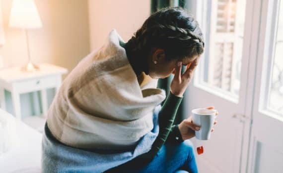 A woman wrapped in a blanket sitting in a hospital room while drinking tea, used to explain COVID-19 and the delta variant