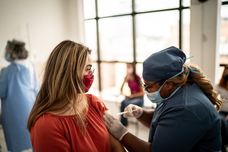 A woman in a red blouse and face mask receiving a vaccination from a medical professional