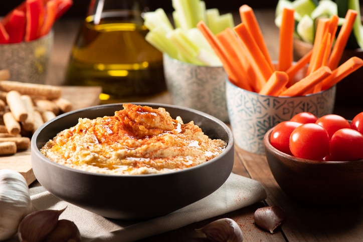 Black bowl with red pepper hummus and raw celery, carrot, and tomato on the side