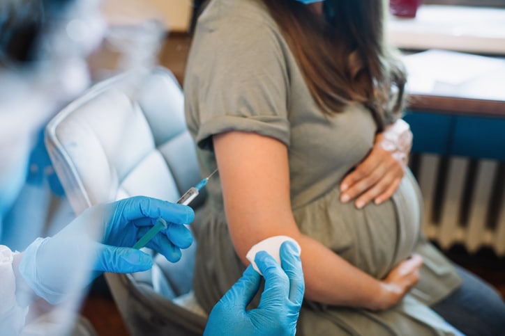 A pregnant woman holding her abdomen, while a medical professional holds a syringe and cotton pad in the foreground