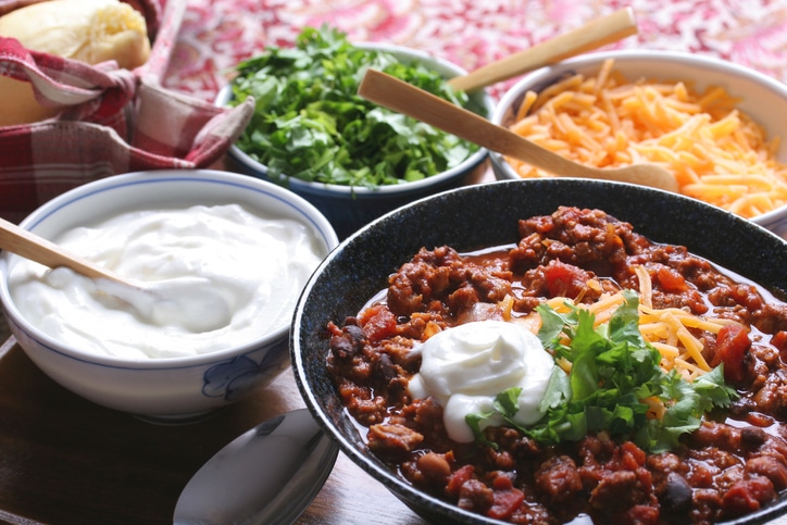 Bowl of beef chili with side bowls of comfort food fixings including sour cream, greens and shredded yellow cheese