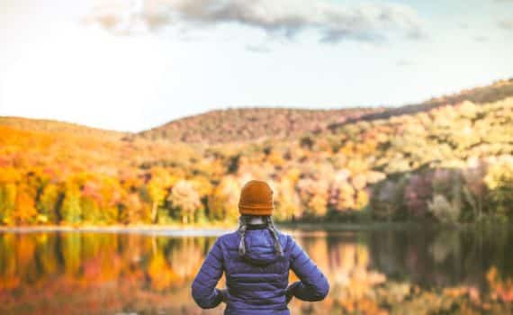 A woman with a red beanie in front of a scenic hilly area with red and yellow trees, used to explain thanksgiving stressors
