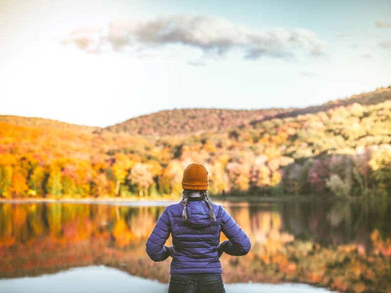 A woman with a red beanie in front of a scenic hilly area with red and yellow trees, used to explain thanksgiving stressors