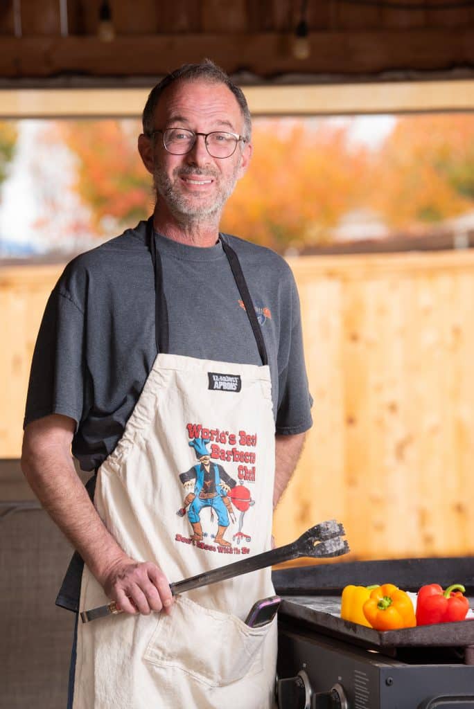 Roger Holcomb holding metal tongs and standing over a grill with bell peppers 