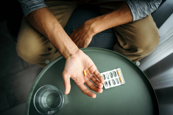A person holding a pill hovers their hand over a table with a glass and pill case on it