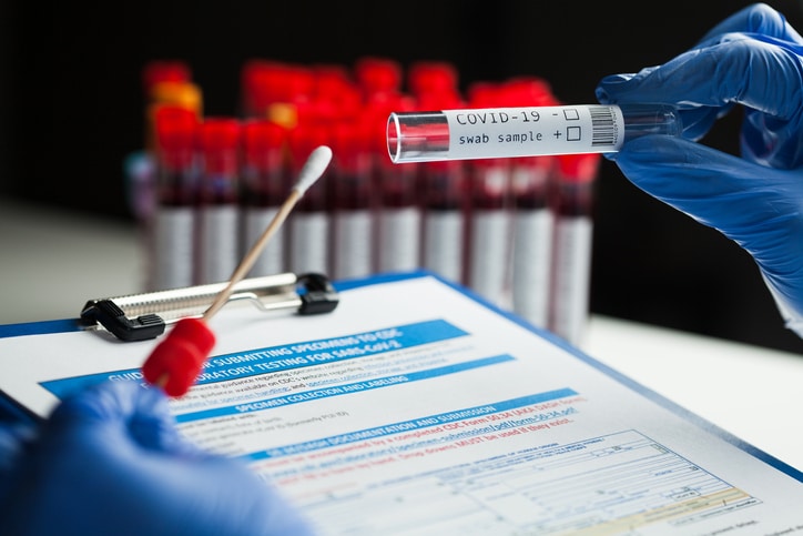 A person wearing gloves holds a vial container that says "COVID-19 swab sample" over a clipboard with documents, with a group of vials with swap samples in the background.