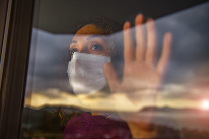 A woman looking outside with a mask on, viewed through a window