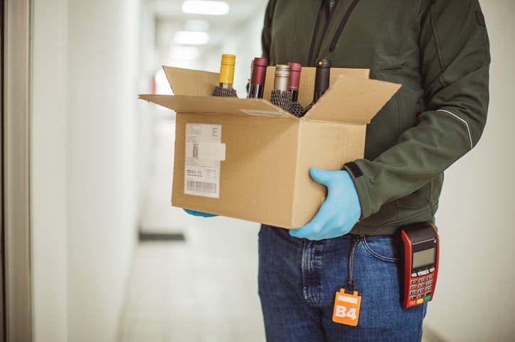 A person with a product scanner on their belt holds a box with at least six wine bottles