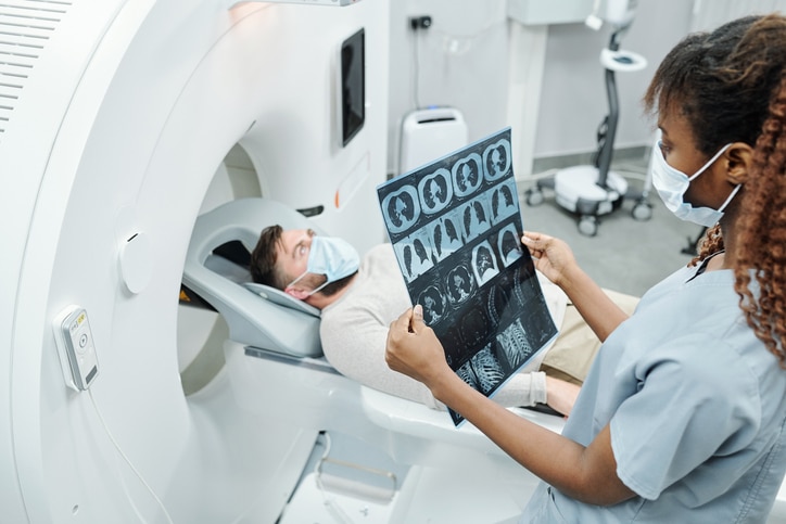 A person sitting on the table of a piece of imaging equipment with a radiologist looking at scans