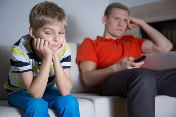 A man holding a television remote and looking off screen, with a child in focus in the front of the image looking to the side with his hands resting on his chin