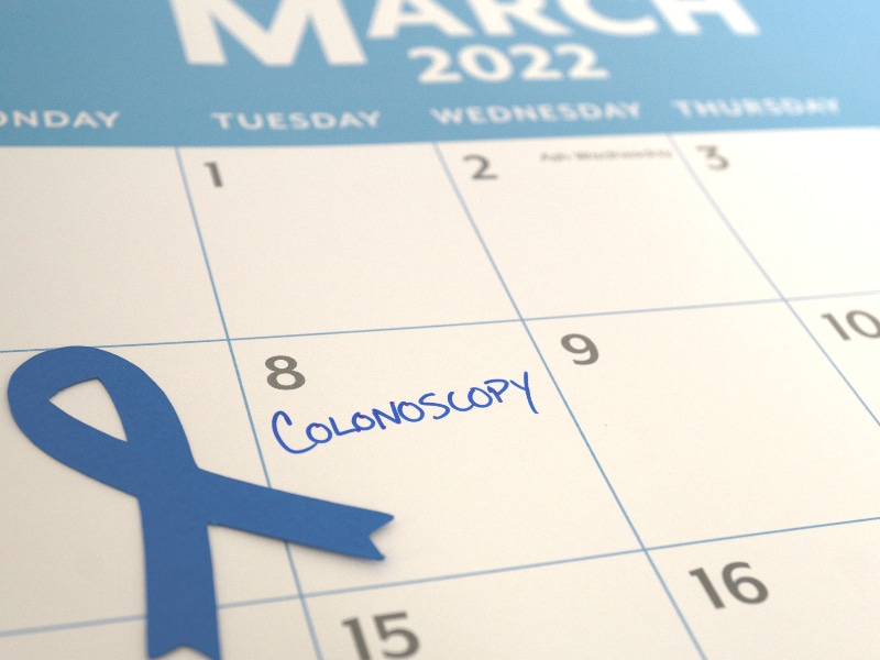 Calendar page of March 2022 open with the word "Colonoscopy" written in blue on the 8th with a blue ribbon next to it, used to explain colon cancer screenings