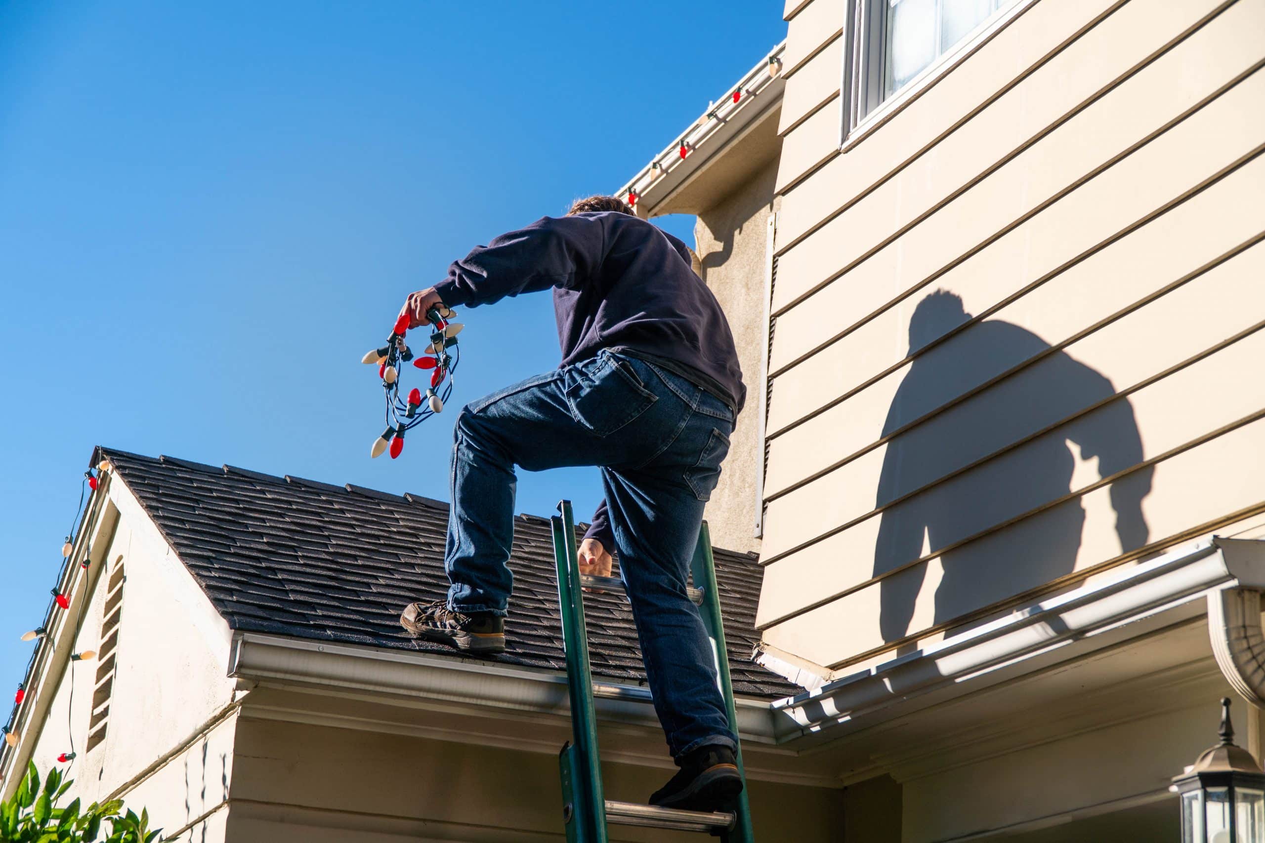 Man climbing ladder onto roof holding exterior colorful lights