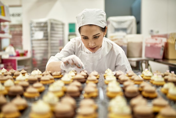 A baker in a white baking coat and cap adds decorations to dozens of cupcakes, her hands placing one decoration on one of the cupcakes