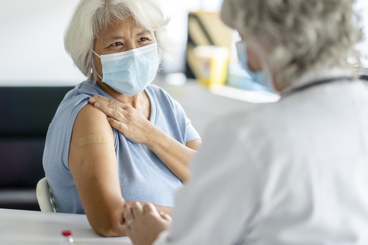 A woman in a medical mask looking to a medical provider in a white coat, pulling up her sleeve to show a bandage on her arm.