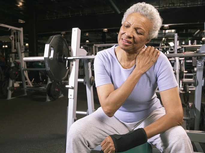 A woman in a gym reaching for her shoulder with a grimaced look on her face