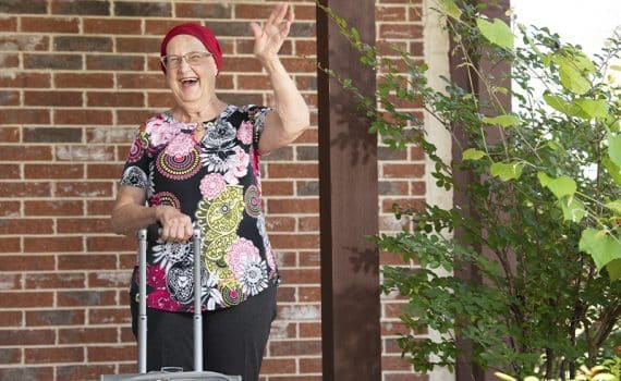 Mary Nash, uterine cancer survivor, wearing a colorful top and holding a suitcase handle, waves happily.