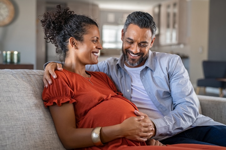 A man and a woman smiling and the man looking down at the woman's baby bump