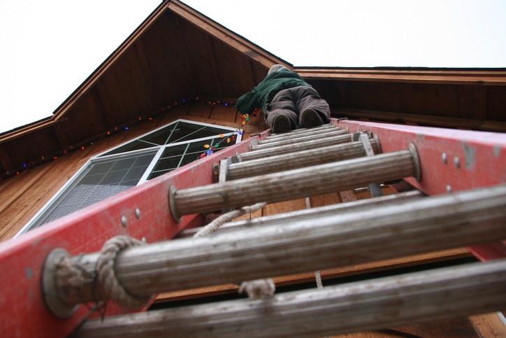A photograph looking up of a person at the top of a ladder while they attach string lights under a roof lining